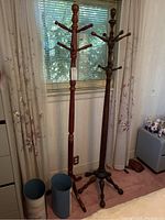 Two wooden coat racks standing side by side near a window, one red-toned and the other dark-toned with multiple hooks each.