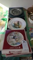 Collector plates including a Royal Albert plate with original box, a colorful bird plate, and a pewter plate in a green box.