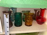 Six miniature crackle glass items on a white shelf including pitchers, vases, and a bowl in amber, red, turquoise, blue, purple, and green colors with a tape measure showing a height about 3 to 7 inches.