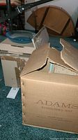 Boxes labeled Adams, vintage dishes in original packaging, including one partially visible tea cup with floral pattern.