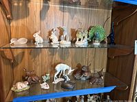 Wide shot showing two shelves with various animal figurines including rabbits, peacock, leopard, giraffe, deer, small bird, dog and flowers.