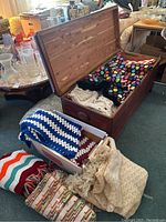 Photo showing multiple handmade afghans in various colors and crochet styles, some folded and some loosely arranged inside and outside a wooden chest.