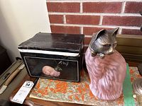 Pair of pink glass body lamps, one with a metallic cat head with a bow tie, displayed next to original box and placed on a wooden surface with orange and white cloth.