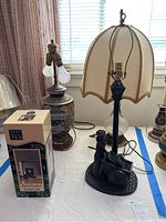 Wide view showing the Asian themed lamp, figurine push cart lamp, and flip-up lamp in box on a table with light curtains background.
