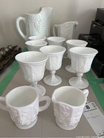 Seven white milk glass goblets and two pitchers with grapevine pattern in the background, one sugar bowl and one creamer in front.