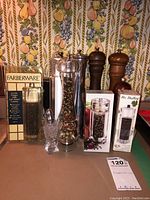 Overview photo showing all peppermills and salt shakers arranged in front of kitchen wallpaper with fruit design, with two boxes and loose salt shakers in front.
