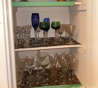Multiple stemware pieces on two cabinet shelves showing clear, green, and blue wine and martini glasses with decorative etched patterns and various stem styles.