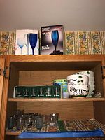 Shelf showing boxed Luminarc and Argold Crystal stemware sets, stacked ceramic bowls with animal and shamrock designs, shot glasses in green plastic rack and additional shot glasses below.
