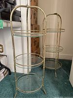 Two 3-tier brass-framed glass-topped tables with rounded edges and bamboo-inspired legs, placed on green carpet near boxes.