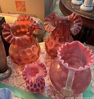 Three pink glass vases and one pink glass pitcher set on a table showing hobnail pattern and ruffled rims.