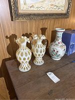 Photo showing three vases on a wooden surface against a wood-paneled wall. Two white art glass vases with gold patterns (one with handles) and one Asian ceramic vase with colorful floral and bird decorations.