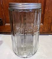 Full view of the 1930s Hoosier coffee canister jar showing ribbed embossed glass and original aluminum lid on white base with wooden background.