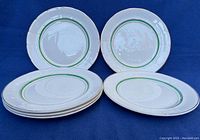 Six white round porcelain plates with green and gold banding around the rims shown against a blue background, arranged standing and stacked for display.