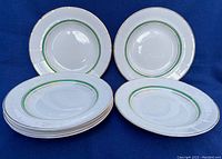 Set of 7 white porcelain plates with green and gold trim arranged in two stacks and two plates displayed on a blue fabric background, showing the overall design and quantity.