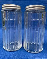 Pair of clear glass antique spice jars with aluminum lids, showing patterned glass sides and screw-on tops.