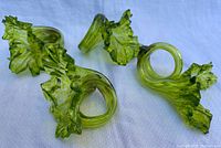 Four green translucent glass napkin rings with leaf and shell-like ruffled designs, arranged on a white textured surface.