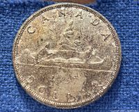 Reverse side of the 1957 Canadian silver dollar coin showing a canoe with two paddlers, dated 1957, with visible tarnishing and wear on the surface, photographed on a blue background.