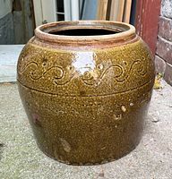 Side and angled views of a large brown glazed stoneware jar with embossed swirl decoration, highlighting its rustic folk pottery style and wide mouth opening.