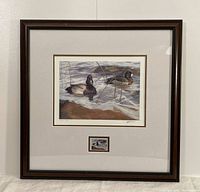 Framed artwork showing a print of two ducks on water with a Canadian stamp of the same image below. Frame is dark brown wood with light mat.