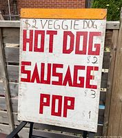 Front view of the large hand-painted wooden sign showing bold red lettered text advertising hot dogs, sausages, veggie dogs, and pop with hand-marked prices.