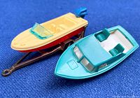 Two vintage plastic boats displayed together on blue background - yellow Matchbox boat with blue windshield; teal/aqua colored Husky Models boat on bronze trailer