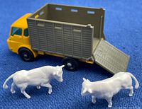 Front and side view of the yellow and gray Matchbox cattle truck displaying the open rear gate with two white plastic cattle in front of the truck on blue background.