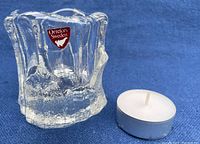 Clear Orrefors Capella crystal candle holder with original Orrefors Sweden sticker placed on front face next to small white tea light candle in metal casing, against blue background.