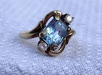 Close-up photo showing the rectangular blue glass center stone and two smaller clear glass stones on a yellow gold band with curved design details.