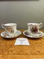 Two Paragon teacup and saucer sets displayed on a wooden surface. Each teacup has a matching saucer. One set has yellow roses, the other has purple and yellow flowers.