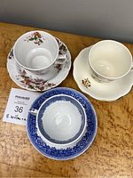 Three ceramic cup and saucer sets displayed together on a wooden surface. Front shows the blue willow pattern cup and saucer in blue and white, with two differently patterned floral cups and saucers behind it.
