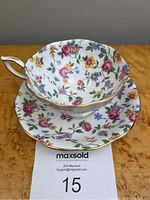 Photo showing the teacup placed on the matching saucer, both decorated with vibrant floral chintz pattern consisting of roses and daffodils with gold trimmed edges, on a wood surface.