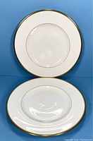 Front and side view of a white porcelain salad plate with a gold rim on a blue background.