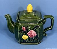 Front side showing floral decoration with pink and yellow raised roses and green leaves, yellow rosebud lid handle, dark green body.