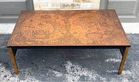 Front angled view of the coffee table showing the copper-toned world map top with hemispheres and decorative map details, metal legs with a gold finish, and black apron.