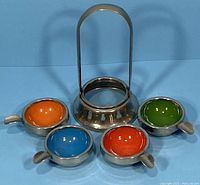 Photo showing all four colorful enameled ashtrays arranged in front of the metal stacking stand with handle against a blue background.