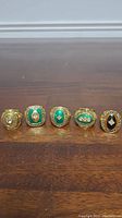 Five Green Bay Packers replica Super Bowl rings arranged on a wooden surface showing front faces with green, black, and clear stones and gold tone metal.