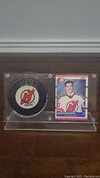 Front view of the clear acrylic display case holding the autographed New Jersey Devils puck and Martin Brodeur rookie card side-by-side on wooden floor.