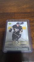 Front view of Sidney Crosby rookie hockey card showing player in black Penguins uniform holding hockey stick, with text and team logo on card.