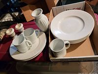 Six stacked white dinner plates in a cardboard box, with additional white ceramic items including mugs, creamers, a vase, and small containers on the side.