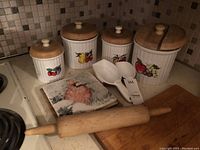 Wooden lid canisters with fruit designs, vintage rolling pin, scoops, and stoneware trivet with bird illustration