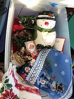 Photo showing a white ceramic snowman cookie jar, blue snowman fabric item, poinsettia-themed white fabric, and some artificial holiday greenery.