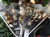Overview photo of various small nativity scenes and Christmas figurines arranged on a glass table, including painted and unpainted ceramic figures, rustic dolls, and decorative ornaments.