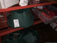 Three green plastic Christmas tree stands with metal tightening hardware on shelving and a measuring stick for scale