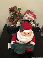 Photo showing plush Santa doll sitting in front of a black bin containing various Christmas decorations like snowmen figurines, decorative baskets including a cornucopia, and gift boxes.