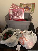 Gray plastic storage tub with Christmas gift boxes, bows, and faux greenery inside. Two plastic shopping bags with more holiday decorations are placed beside the tub.