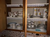 Wood cabinet shelf with various dishware, including mugs with floral and scenic designs, stacks of plates, bowls, creamer, and cups.