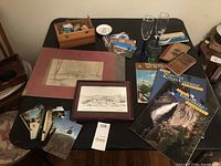 Photo showing table with framed sketches, travel magazines, postcards, commemorative glasses, wooden box with ephemera, and assorted printed materials
