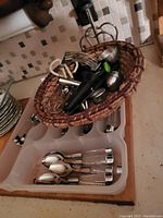 Flatware spoons and forks with two different patterns placed inside a wicker basket and plastic drawer organizer, also showing additional handheld kitchen utensils such as a black handheld mixer and potato masher