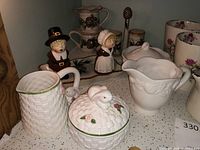 Shelf display of vintage serveware including salt and pepper shakers shaped as pilgrims, bunny-themed sugar bowl, white basket-weave creamer, and floral mugs on a shelf with lace trim.