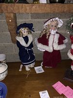Two porcelain holiday dolls on display, one in navy blue velvet dress with white fur, the other in red velvet cloak with white fur.